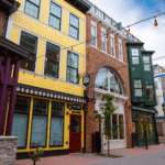 A street scene showcasing a row of colorful buildings, including a yellow building with fish scale siding, a brick building with an arched window, and a green building with large windows. The paved street features sleek Premium Electric vehicles quietly parked along the curb.