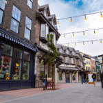 A pedestrian street with buildings featuring shops and cafes, string lights overhead, and a few people walking. The storefronts display various items, creating a quaint, inviting atmosphere. Among the charming shops is a premium electric store showcasing the latest gadgets.