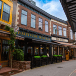 A street view of a wine bar called "Fortitude Wine Bar" and a store named "Wee Shoppe" in a brick building with a yellow awning, outdoor seating area, and premium electric lights illuminating the evening.