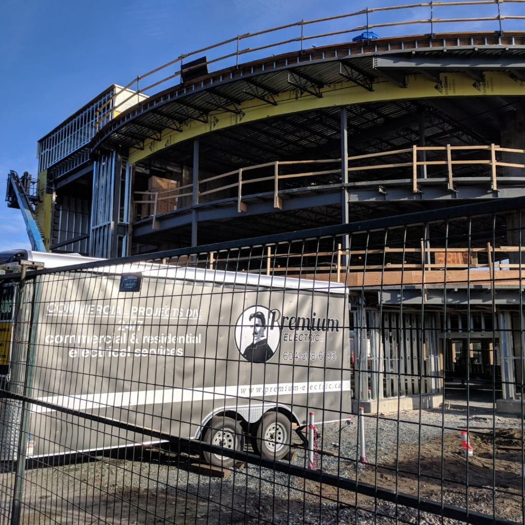 A construction site with a partially completed building and a "Premium Electric" trailer parked nearby. Metal scaffolding is visible, surrounded by a fence.