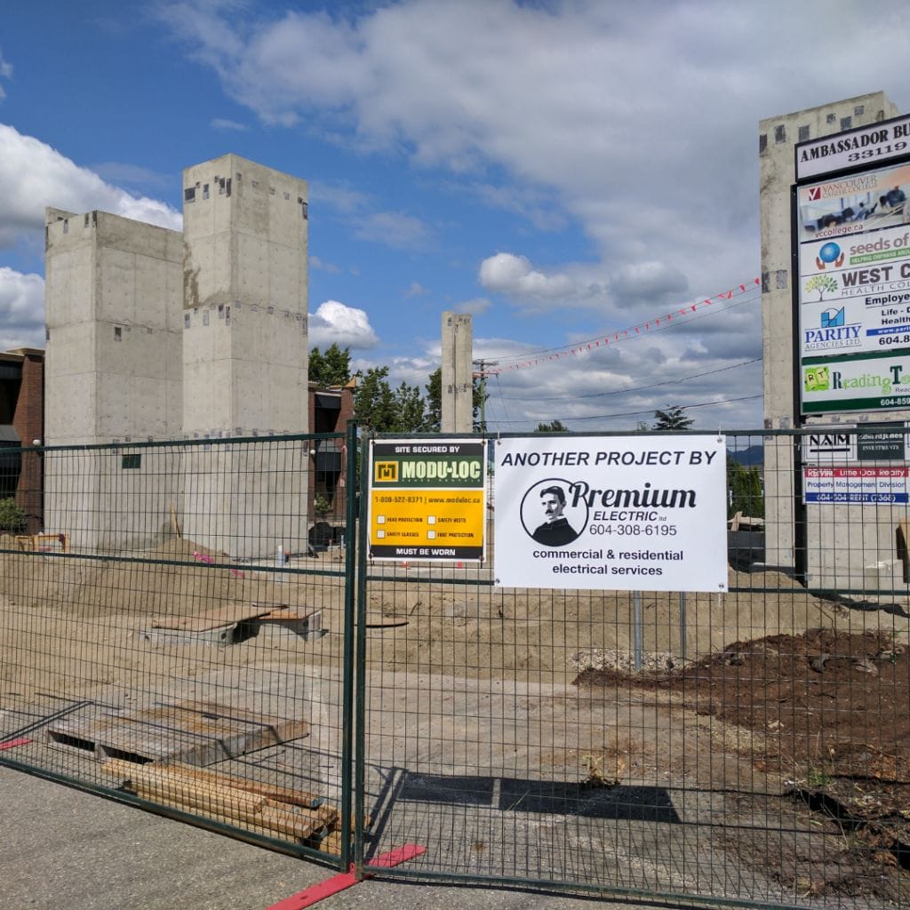 A construction site with concrete pillars and fencing. A sign on the fence reads "ANOTHER PROJECT BY PREMIUM ELECTRIC" alongside another promoting fencing services by Premium Electric.