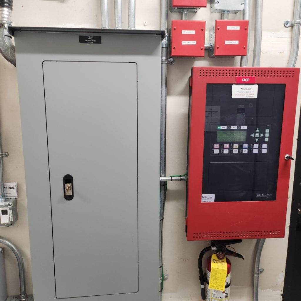 Image of a Premium Electric panel and a fire alarm control panel mounted on a wall. A fire extinguisher is placed below the panels.