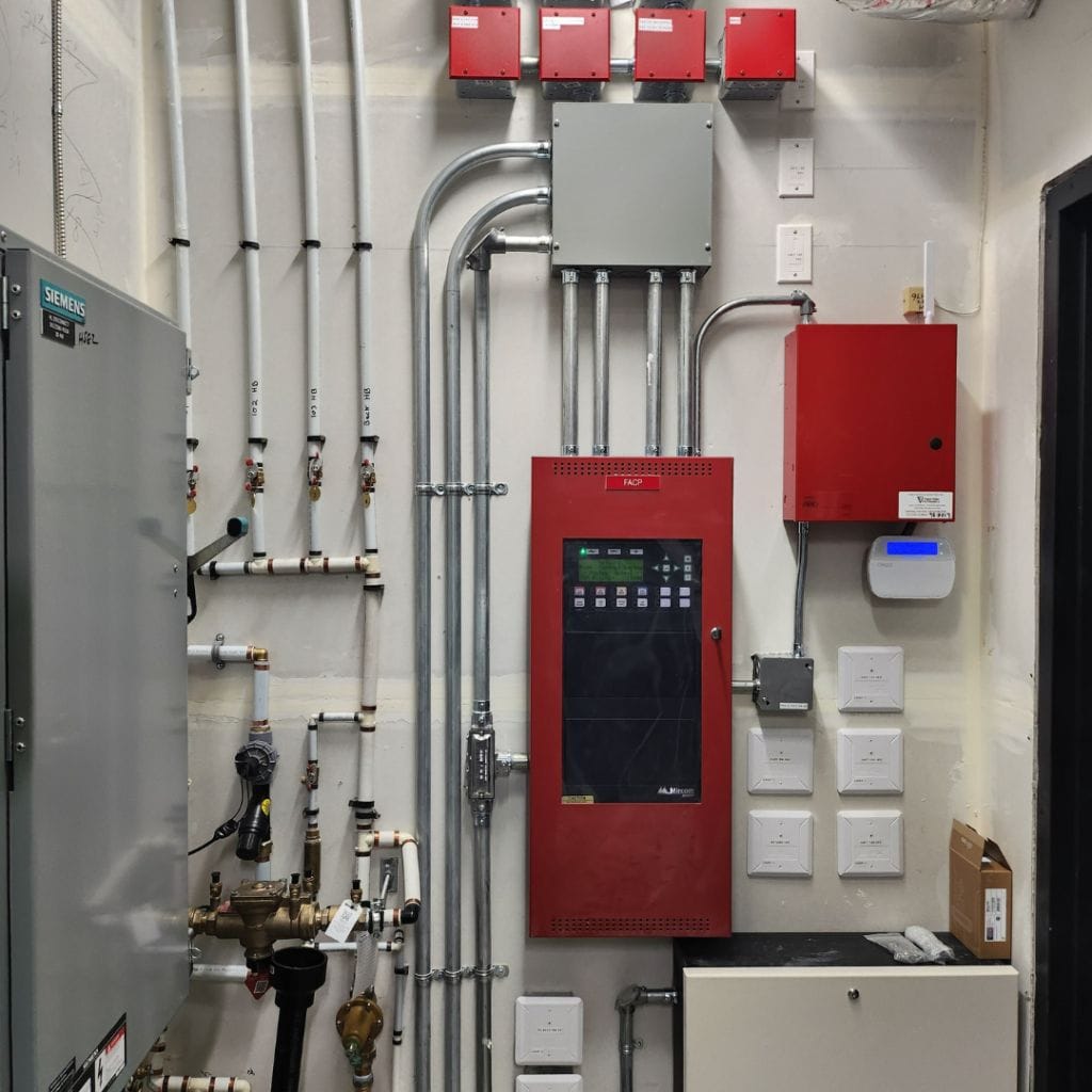 A room with Premium Electric panels, pipes, and a red control unit with a touch screen display mounted on the wall. Multiple cables and conduits run along the wall.