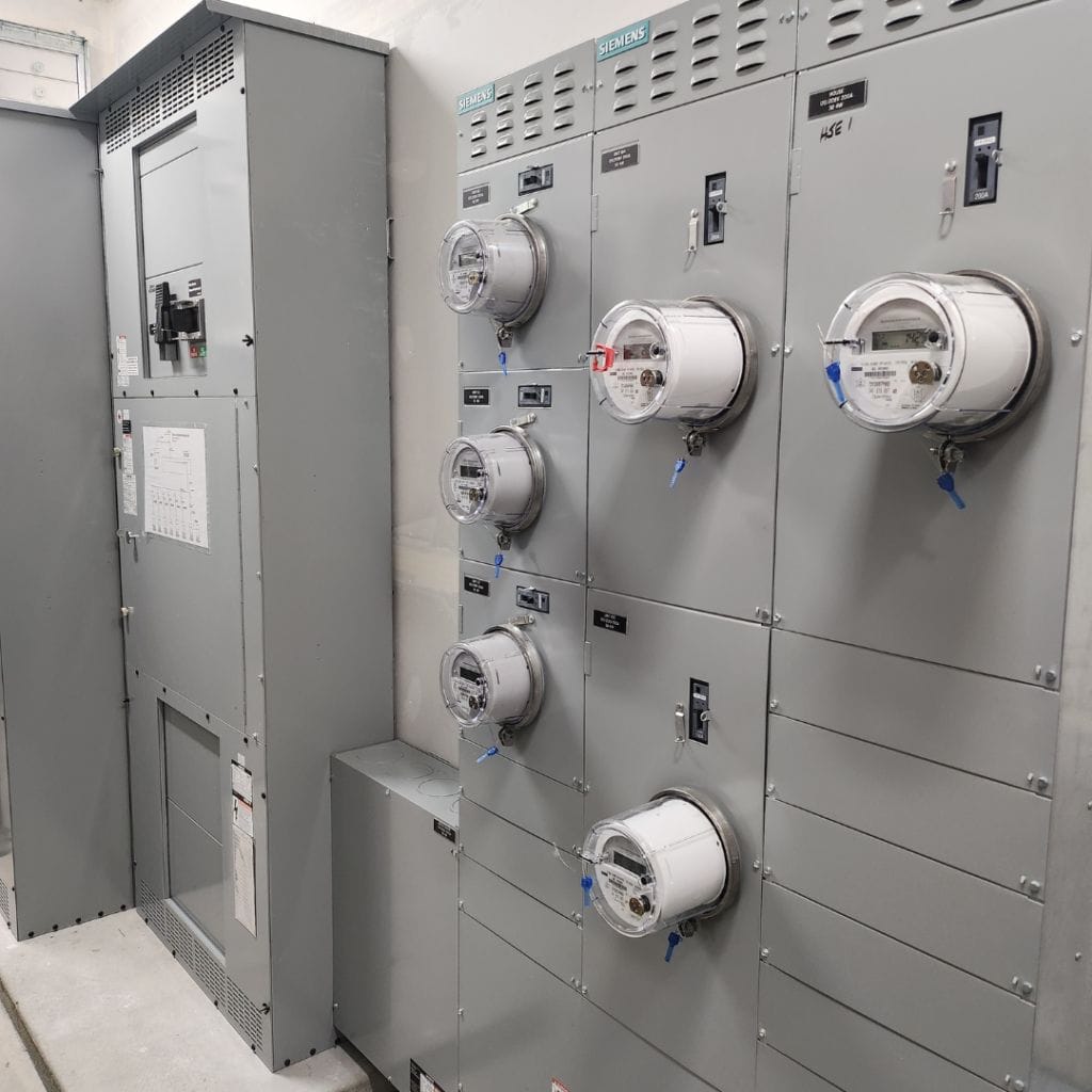 A row of Premium Electric utility meters is mounted on a metal panel adjacent to a large electrical control panel in an industrial setting.