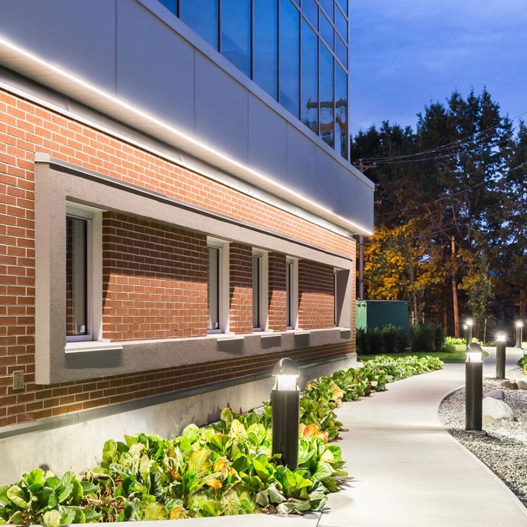 Modern building exterior with brick walls, large windows, and pathway lighting. The landscaped pathway is lined with greenery and illuminated by tall outdoor lamps from Premium Electric, with trees visible in the background.