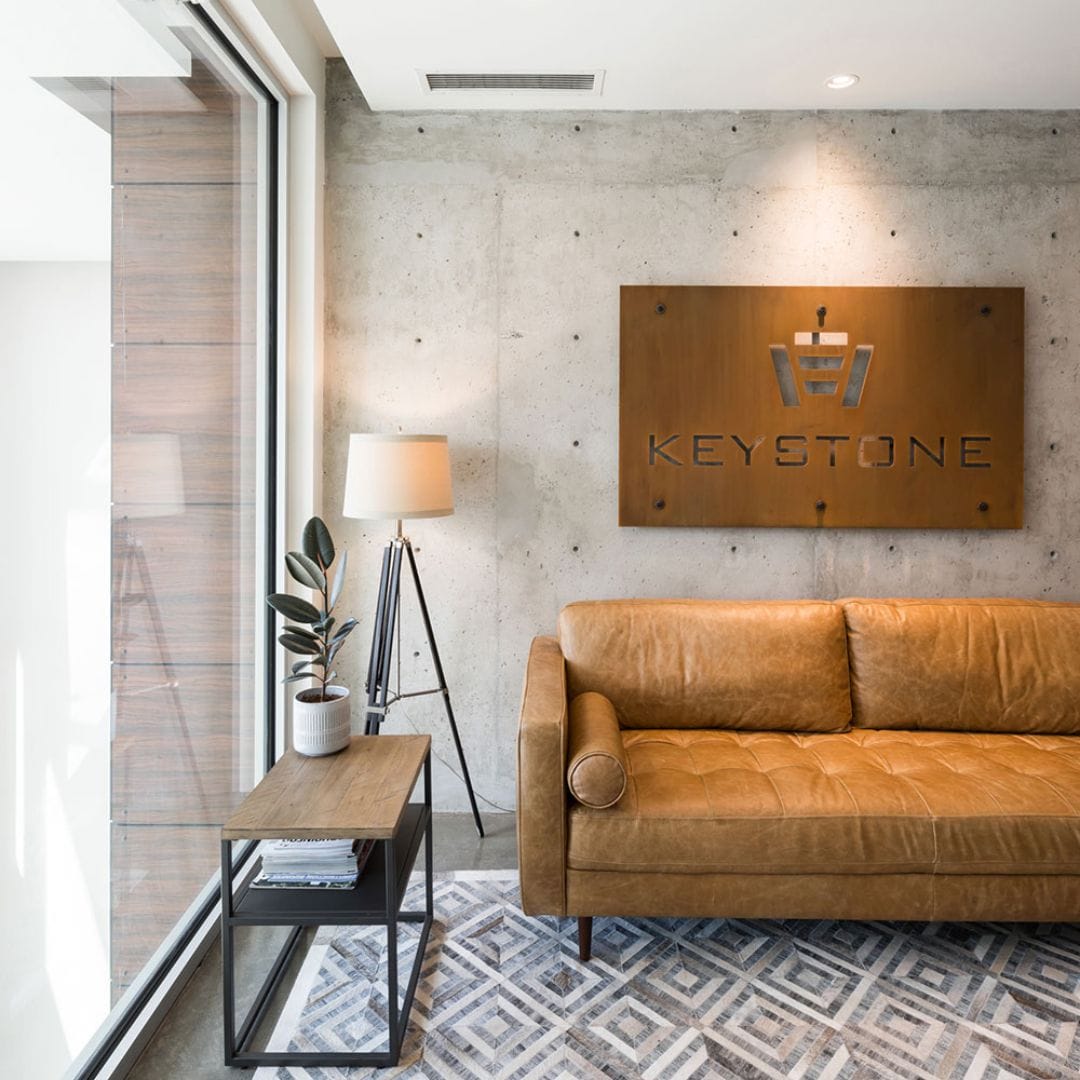 A modern office waiting area features a brown leather sofa, a potted plant on a side table, and a floor lamp. A sign reading "Keystone Premium Electric" is elegantly mounted on the concrete wall.