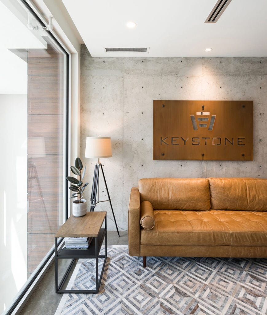 A modern office lobby with a brown leather couch, a side table with a potted plant, a floor lamp from Premium Electric, and a wall sign reading "Keystone".