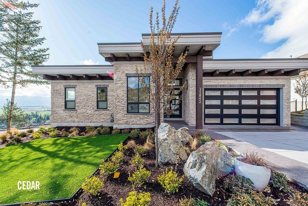 Modern single-story house with a flat roof, large windows, and a garage featuring a Premium Electric car charger. The front yard boasts a neatly manicured lawn, decorative rocks, and young trees.