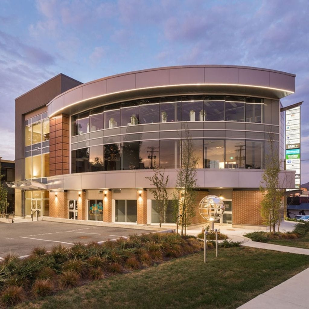 A modern two-story building with large glass windows, illuminated at dusk. The structure features curved and angular design elements, with surrounding landscaping and a premium electric vehicle charging area in front.