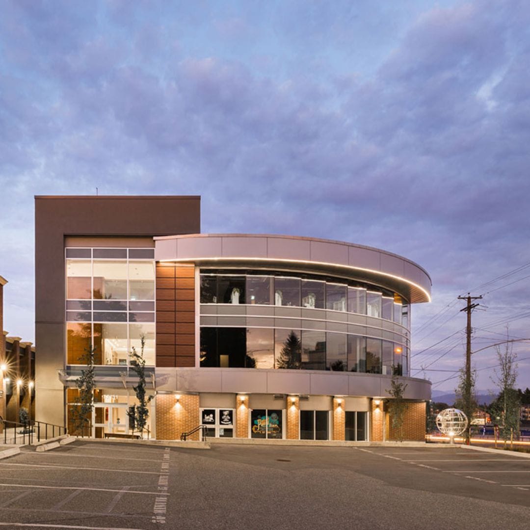 A modern, round, multi-story building with large windows and an adjacent parking lot, featuring premium electric vehicle charging stations, set against a cloudy sky at dusk.