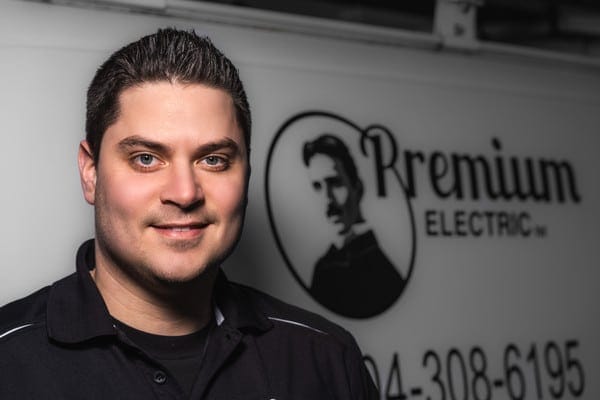 A man in a black shirt stands in front of the "Premium Electric" sign, featuring a logo of a mustachioed man. A partial phone number is visible.