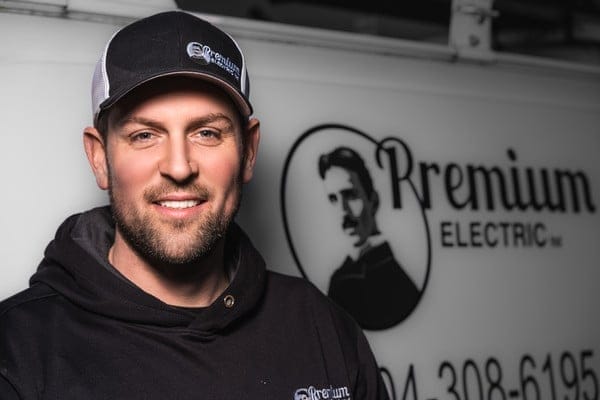 A man in a black baseball cap and hoodie branded with "Premium Electric" stands confidently by a vehicle showcasing the same logo and contact number.