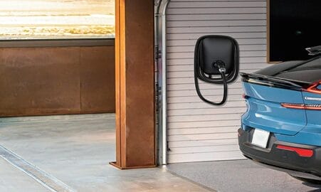 Blue electric car in a garage, connected to a wall-mounted charging station.