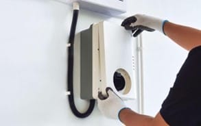 A person wearing gloves installs or inspects a wall-mounted device with wires, setting up an EV charging station at home.