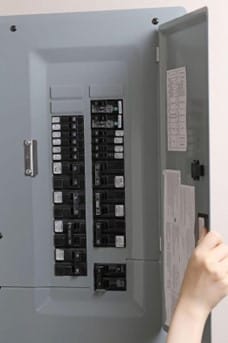 A person opens the door of a gray electrical breaker panel, revealing multiple switches tailored for seamless EV charging at home.