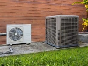 Two outdoor air conditioning units are placed on concrete pads next to a wooden wall, with grass in the foreground—a common sight near homes where knob and tube wiring FAQs are often discussed.